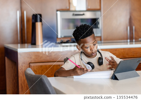 African American boy writing with pencil in notebook at kitchen table, gazing at tablet, copy space 135534476