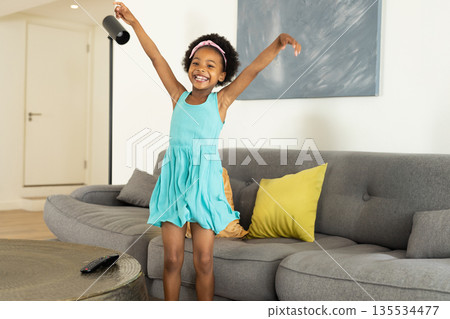 Girl child standing on gray sofa in living room, raising arms with TV remote on table Girl child standing on gray sofa in living room, raising arms with TV remote on table 135534477