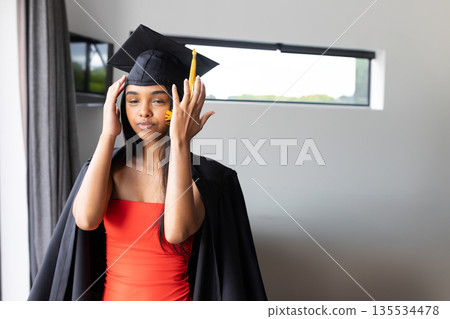 Woman adjusting mortarboard cap indoors in modern room, wearing graduation gown over red dress Woman adjusting mortarboard cap indoors in modern room, wearing graduation gown over red dress 135534478