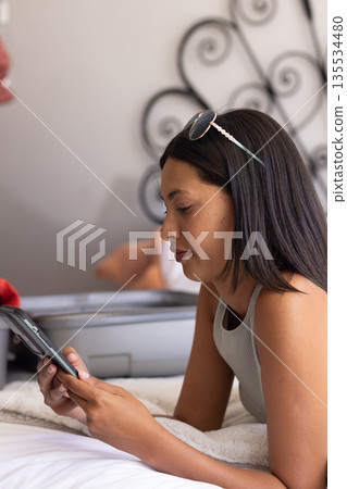 Young multiracial woman lying on bed using smartphone in bedroom with grey suitcase, copy space 135534480