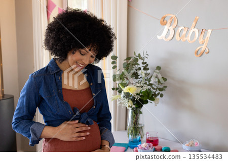 African American woman standing in dining area cradling bump near table with macarons, baby banner 135534483