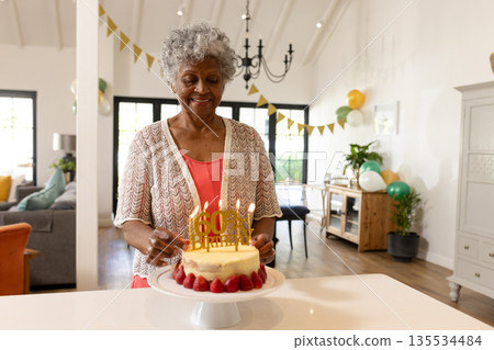 Senior woman adjusting birthday candles on cake in open-plan living room, with balloons and banner 135534484