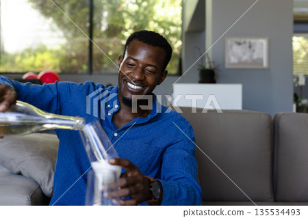 African American man pouring water into glass from pitcher in living room, copy space 135534493