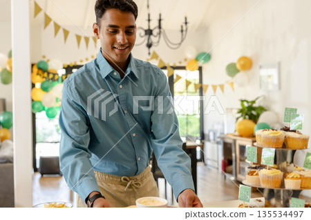 Asian Indian man arranging cups on table in living room, with balloons and cupcakes on stand Asian Indian man arranging cups on table in living room, with balloons and cupcakes on stand 135534497