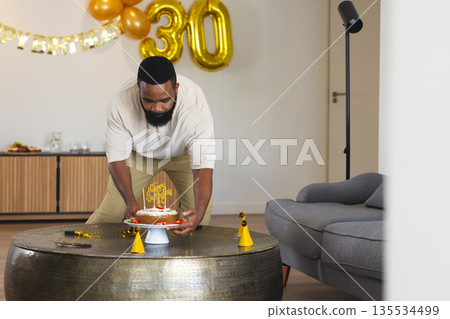 African American man placing birthday cake on coffee table in living room, with gold foil balloons African American man placing birthday cake on coffee table in living room, with gold foil balloons 135534499