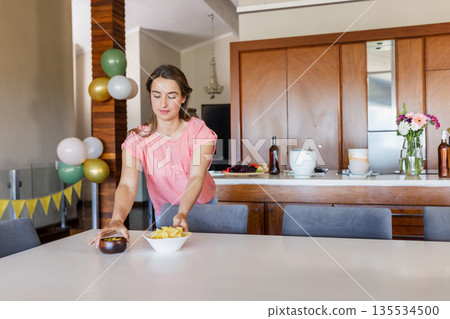 Young woman arranging chips in bowl on dining table in open-plan kitchen, with balloon decorations 135534500