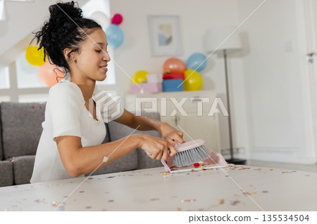 Teenage girl sweeping multicolored confetti off white table in living room, using pink dustpan Teenage girl sweeping multicolored confetti off white table in living room, using pink dustpan 135534504