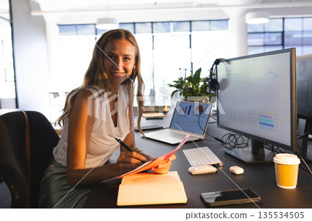 Mid adult woman sitting at desk near large windows, reviewing papers by laptop and monitor Mid adult woman sitting at desk near large windows, reviewing papers by laptop and monitor 135534505
