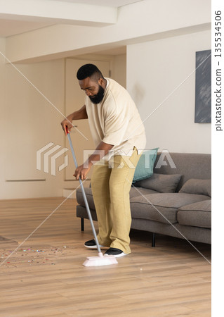 African American man sweeping colorful confetti off light wood floor in living room, with broom 135534506