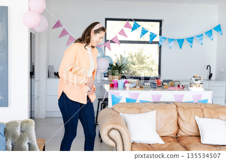 Pregnant woman cradling bump by table with pastel bunting and pink balloons at home, copy space 135534507