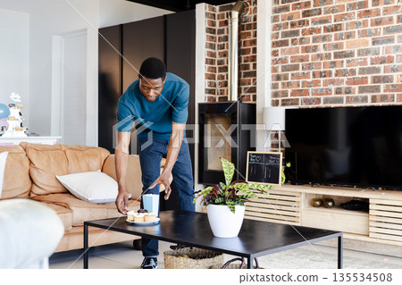 African American man arranging snacks and popcorn container on coffee table in modern living room African American man arranging snacks and popcorn container on coffee table in modern living room 135534508