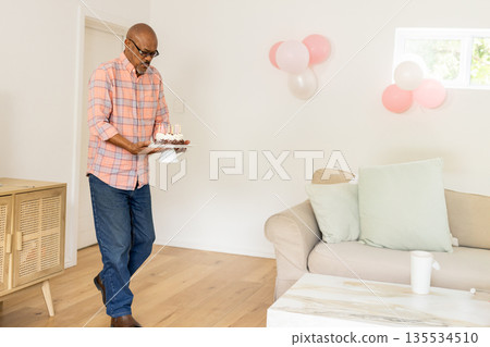Diverse senior man carrying birthday cake on stand in bright living room, with pastel balloons Diverse senior man carrying birthday cake on stand in bright living room, with pastel balloons 135534510