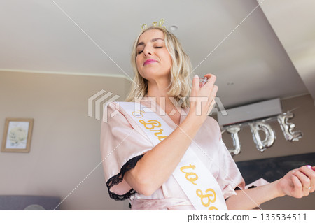 woman spraying perfume from glass atomizer at home, wearing robe and Bride to Be sash woman spraying perfume from glass atomizer at home, wearing robe and Bride to Be sash 135534511