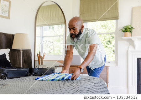 Senior African American man folding striped shirt into black suitcase in bedroom, copy space 135534512
