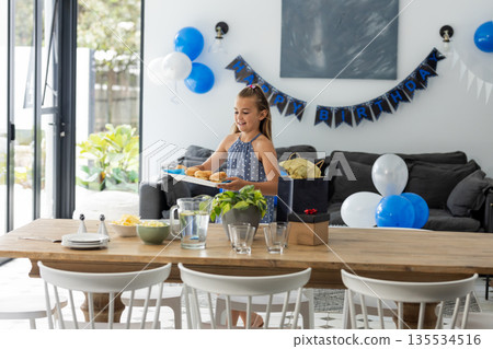 Girl carrying tray of sandwiches across dining table at home with balloons and birthday gifts Girl carrying tray of sandwiches across dining table at home with balloons and birthday gifts 135534516