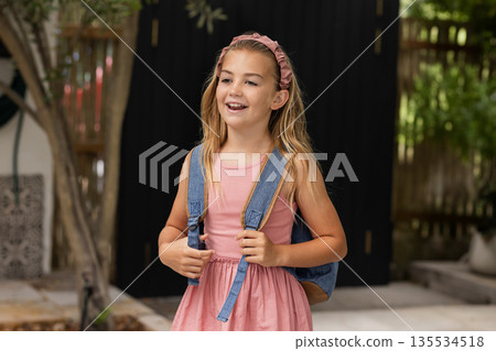 Girl child standing on home patio, holding denim backpack straps, wearing pink headband, smiling Girl child standing on home patio, holding denim backpack straps, wearing pink headband, smiling 135534518