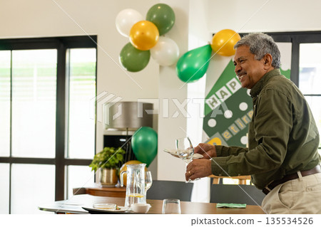 Holding wine glasses middle aged man arranging tableware in dining room, with balloons and banner 135534526