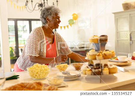 Arranging Senior woman placing cupcakes on kitchen island at home, with balloons and bunting 135534527