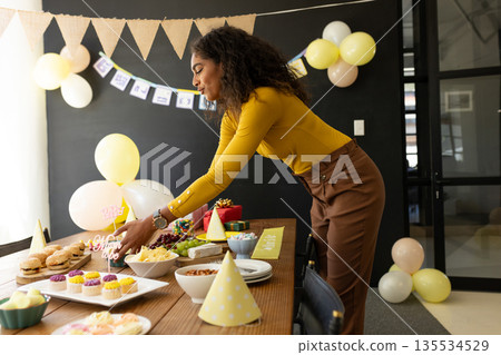 Arranging African American woman leaning over table in office, cake topper and balloons, copy space 135534529