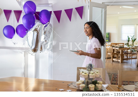 Child girl standing beside table at home holding tablet amid cupcakes and balloons, copy space 135534531