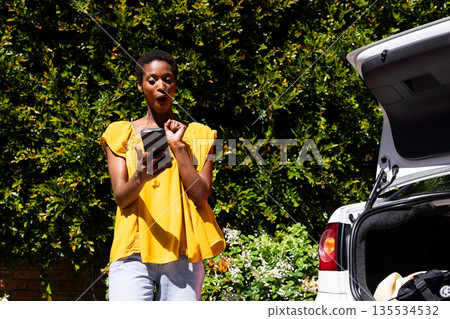 30s African American woman posing beside open car trunk with bag in driveway holding smartphone 135534532