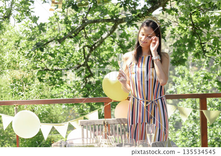 Standing woman wearing tiara and holding champagne flute on deck, with pastel balloons and bunting Standing woman wearing tiara and holding champagne flute on deck, with pastel balloons and bunting 135534545