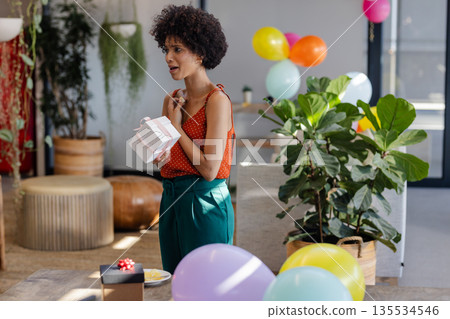 African American female standing in lounge area holding gift box with ribbon amid plants, balloons 135534546