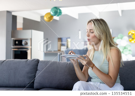 Woman in sleeveless top eating cake by teal gold balloons in modern kitchen area, copy space 135534551