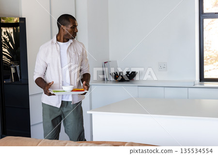 African American man holding white tray with bowl and sliced vegetables in kitchen, copy space African American man holding white tray with bowl and sliced vegetables in kitchen, copy space 135534554