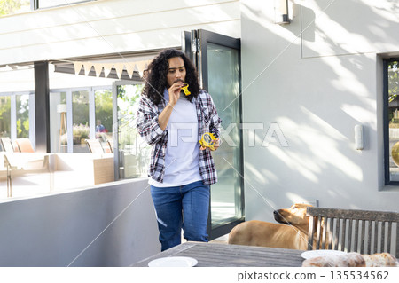 Man stepping onto sunny patio carrying yellow cookie packet and biting cookie next to brown dog 135534562