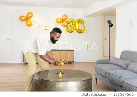 African American man bending at coffee table placing yellow hat and gold 30 balloons, copy space 135534566