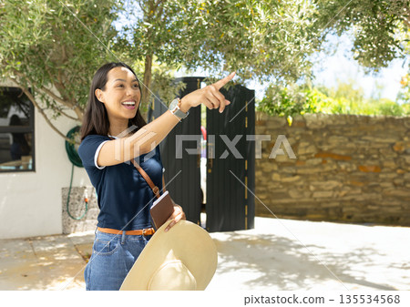 Mid adult Asian woman pointing in sunlit courtyard holding straw hat journal bag and wristwatch Mid adult Asian woman pointing in sunlit courtyard holding straw hat journal bag and wristwatch 135534568