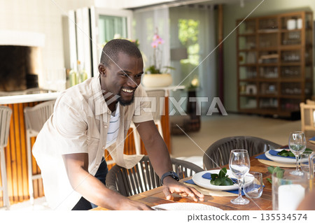 Smiling and leaning across wooden dining table at home with plates, blue napkins and leaf sprigs 135534574