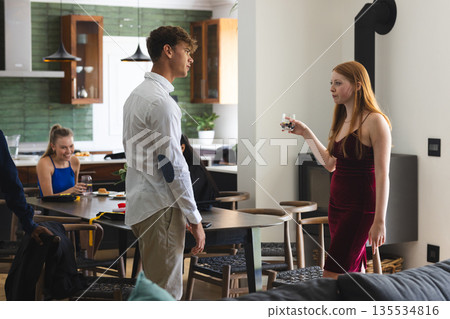Teen girl in red dress holding drink, talking to boy at home celebration Teen girl in red dress holding drink, talking to boy at home celebration 135534816