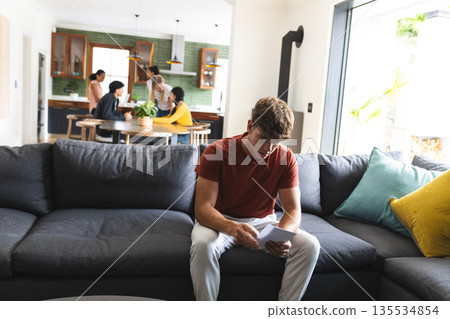Teen reading letter on couch while friends gather around kitchen table 135534854