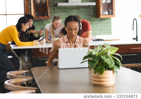 African American woman using laptop at home while teens socializing in kitchen African American woman using laptop at home while teens socializing in kitchen 135534871