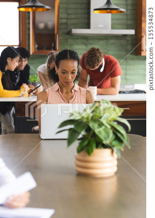 Woman using laptop at home while teens socializing in kitchen background Woman using laptop at home while teens socializing in kitchen background 135534873