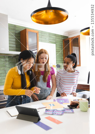 In kitchen, teen girls using tablet and colorful papers for creative project 135534932