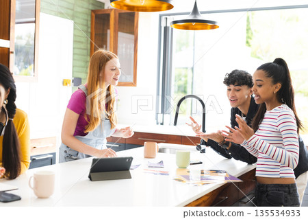 Teen friends in kitchen laughing and chatting with tablet and coffee mugs Teen friends in kitchen laughing and chatting with tablet and coffee mugs 135534933