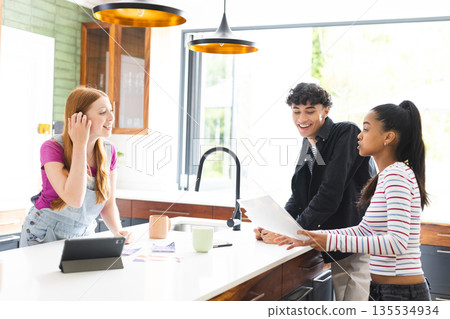 Teen friends in kitchen laughing and discussing project with tablet and papers Teen friends in kitchen laughing and discussing project with tablet and papers 135534934