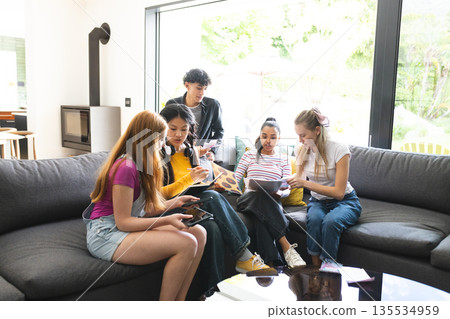 Teen friends studying together on couch, sharing notes and using tablet at home 135534959