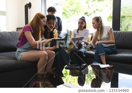 Teen friends enjoying tablet and laptop together on sofa in bright living room 135534960