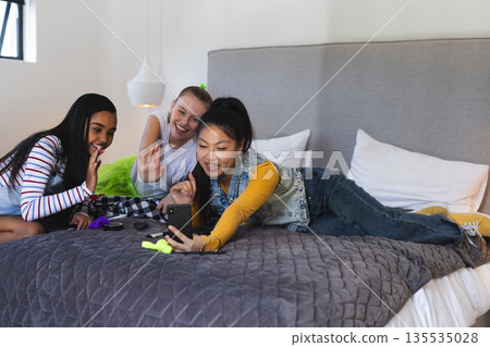 Teen girls video chatting on smartphone, laughing and waving on bed at home Teen girls video chatting on smartphone, laughing and waving on bed at home 135535028