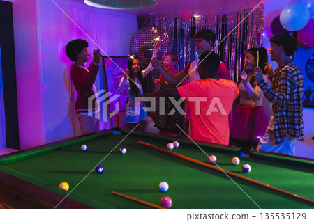 Teenagers celebrating around pool table with colorful lights and festive decorations Teenagers celebrating around pool table with colorful lights and festive decorations 135535129