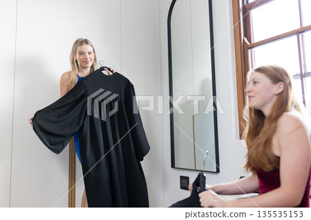 Teen girls excitedly preparing for graduation, holding gowns in bedroom 135535153