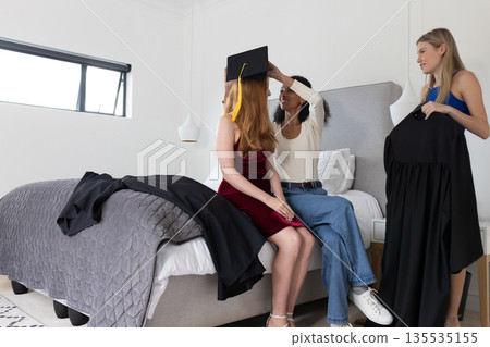 Friends preparing for graduation, adjusting cap and gown in bedroom Friends preparing for graduation, adjusting cap and gown in bedroom 135535155