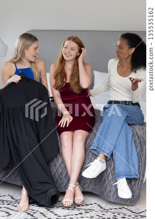 Teen girls discussing graduation gown while sitting on bed, sharing excitement 135535162