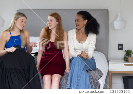 Teen girls discussing graduation outfit choices while sitting on bed at home 135535163