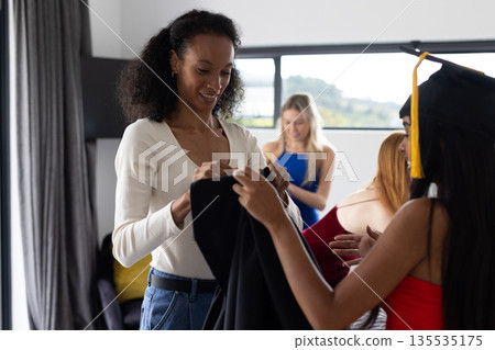 Teen celebrating graduation at home, friends helping with cap and gown 135535175