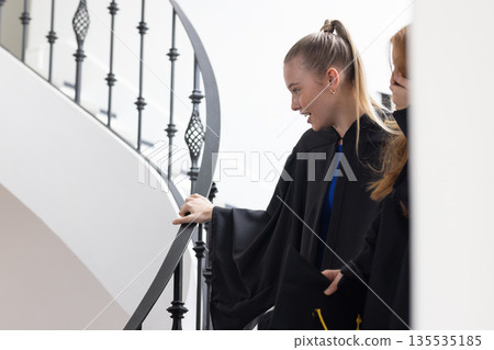 Teen girls in graduation gowns smiling and chatting on staircase indoors Teen girls in graduation gowns smiling and chatting on staircase indoors 135535185